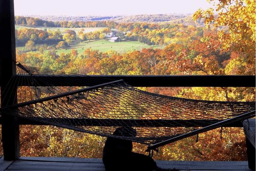 A hammock is draped over a railing with a scenic view of vibrant autumn foliage in the background.